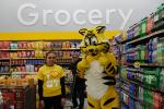 A female Giant Tiger Saint John, N.B. employee posing with Friendly, the Giant Tiger in front of a wall that says the word “Grocery”. 
